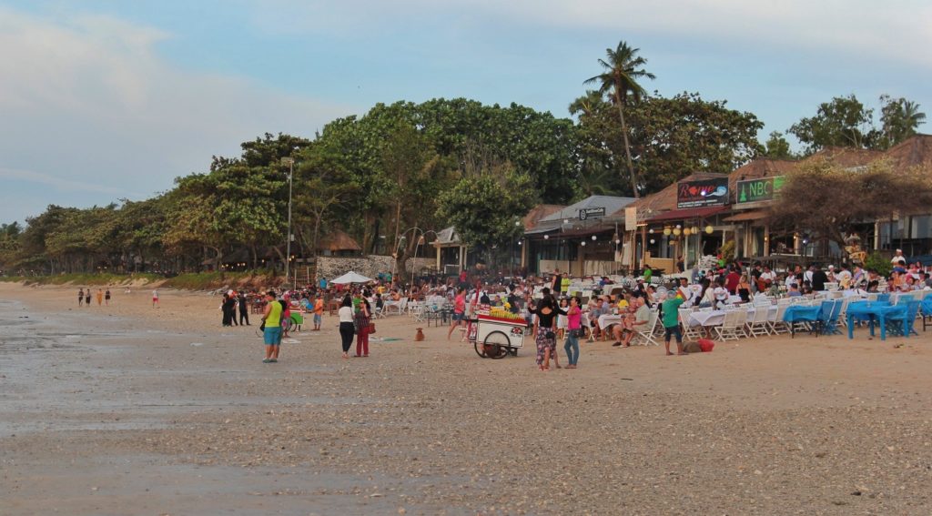 Ein paar Hütten am Rand, viele Tischen mit Besuchern, der Strand ist viel besucht. Im Hintergrund sind Bäume zu sehen.